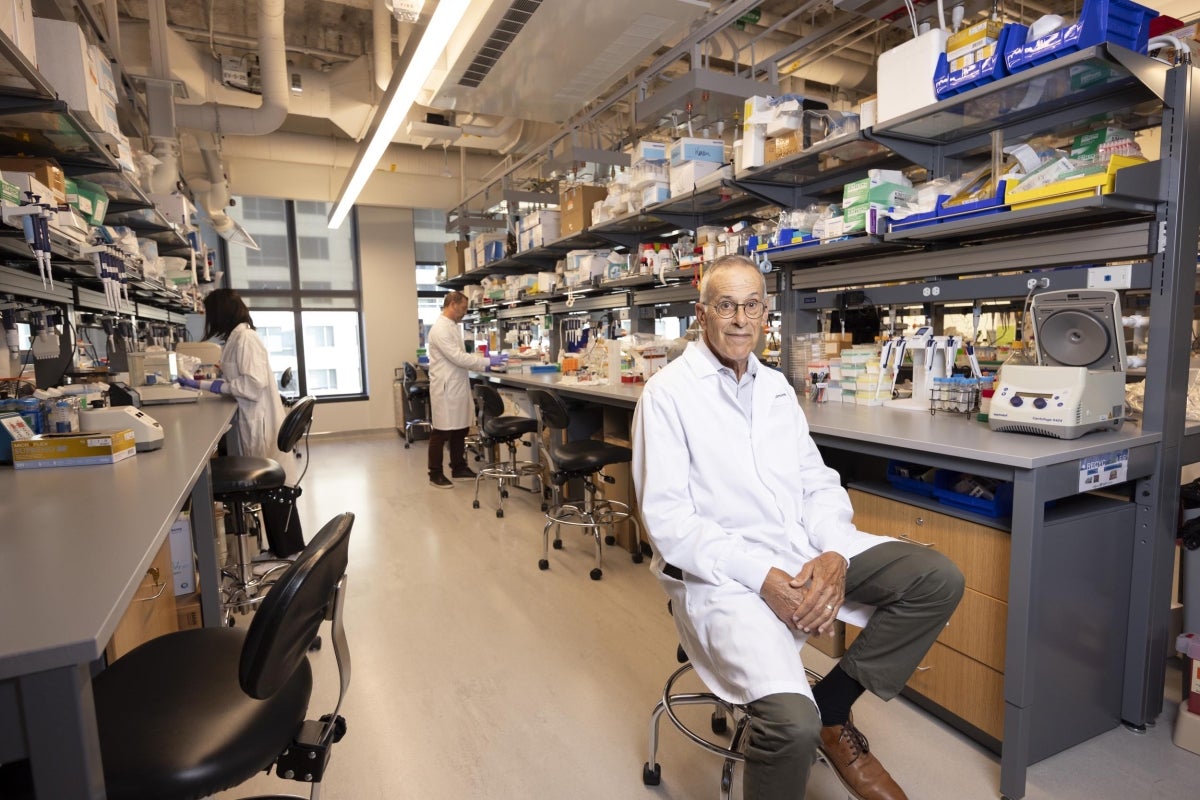 David Walt on a stool in the foreground of a modern biomedical research laboratory, looking at the camera. Behind him, two other researchers in lab coats are working at laboratory benches filled with various scientific equipment, containers, and supplies. The lab has high ceilings with exposed ducts, bright lighting, and large windows letting in natural light.
