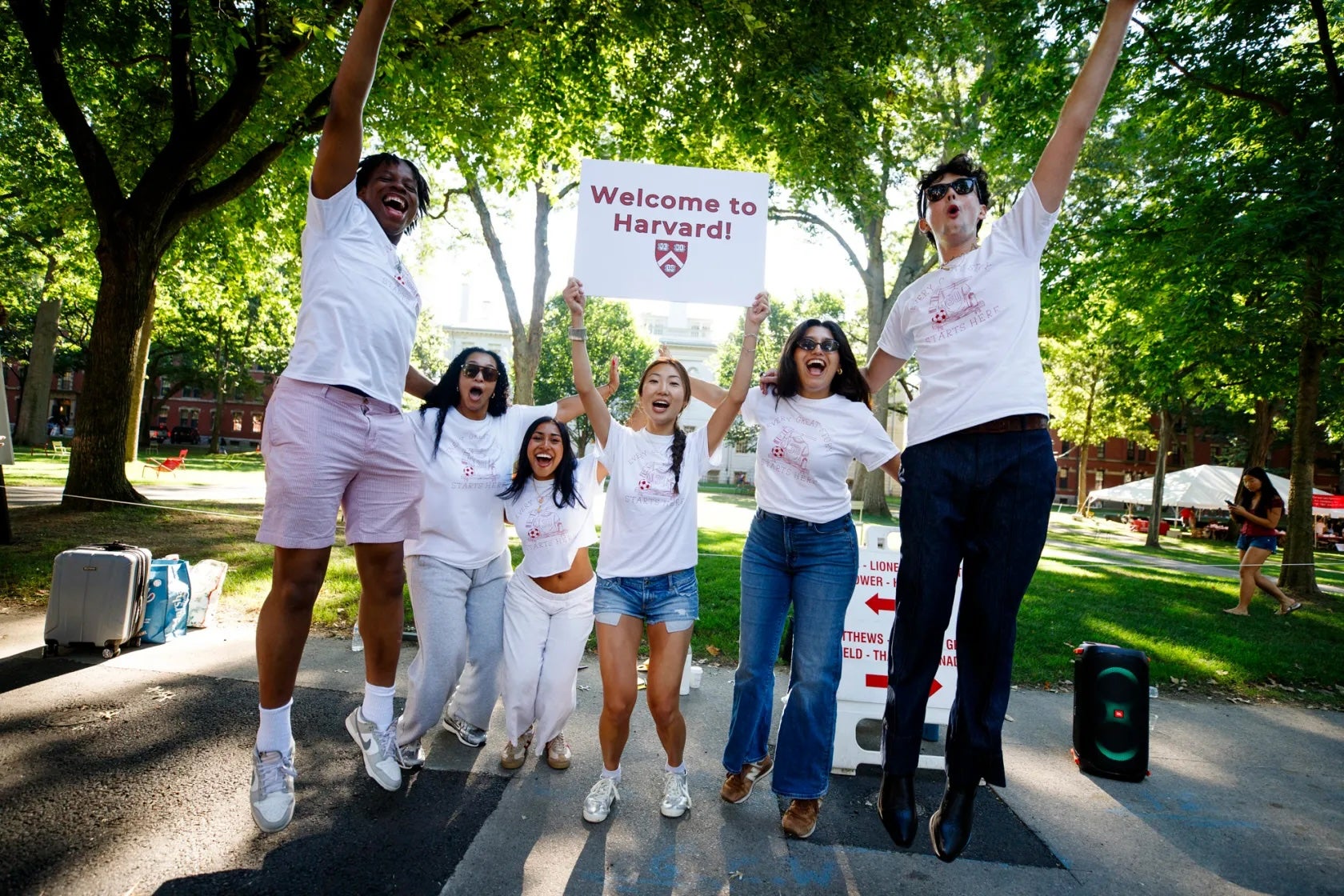 Several students jump and cheer in Harvard Yard, with one holding a Welcome to Harvard! sign.