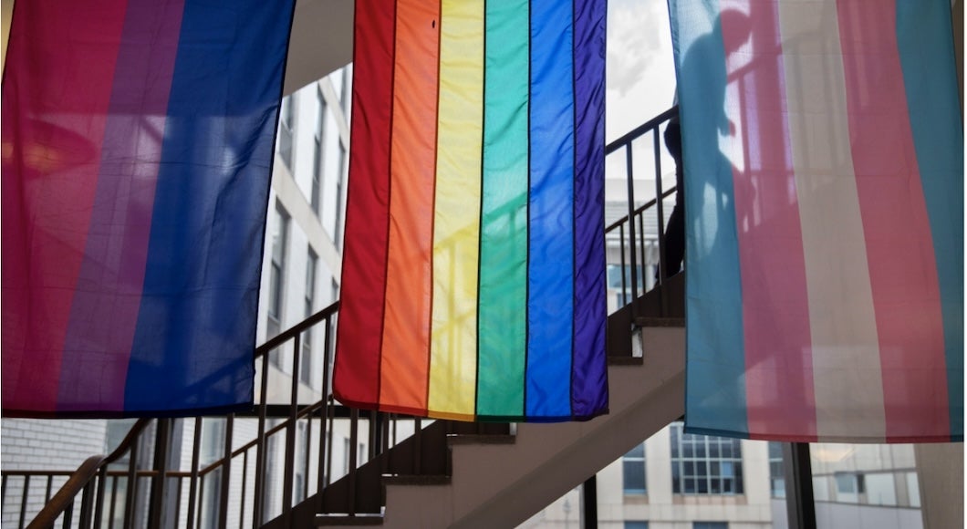 Three LGBTQ identity pride flags (a bisexual pride flag, a rainbow LGBTQ pride flag, and a transgender pride flag) hang from the ceiling inside the Harvard Chan School. In the background behind the flags, a person climbs a staircase.