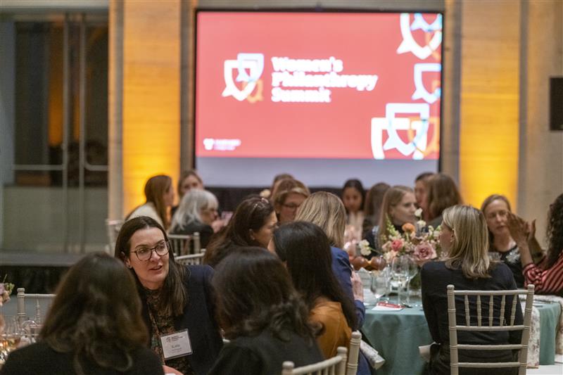 Large banquet-style room filled with round tables where women are seated and talking; a big screen at the front reads “Women’s Philanthropy Summit.”