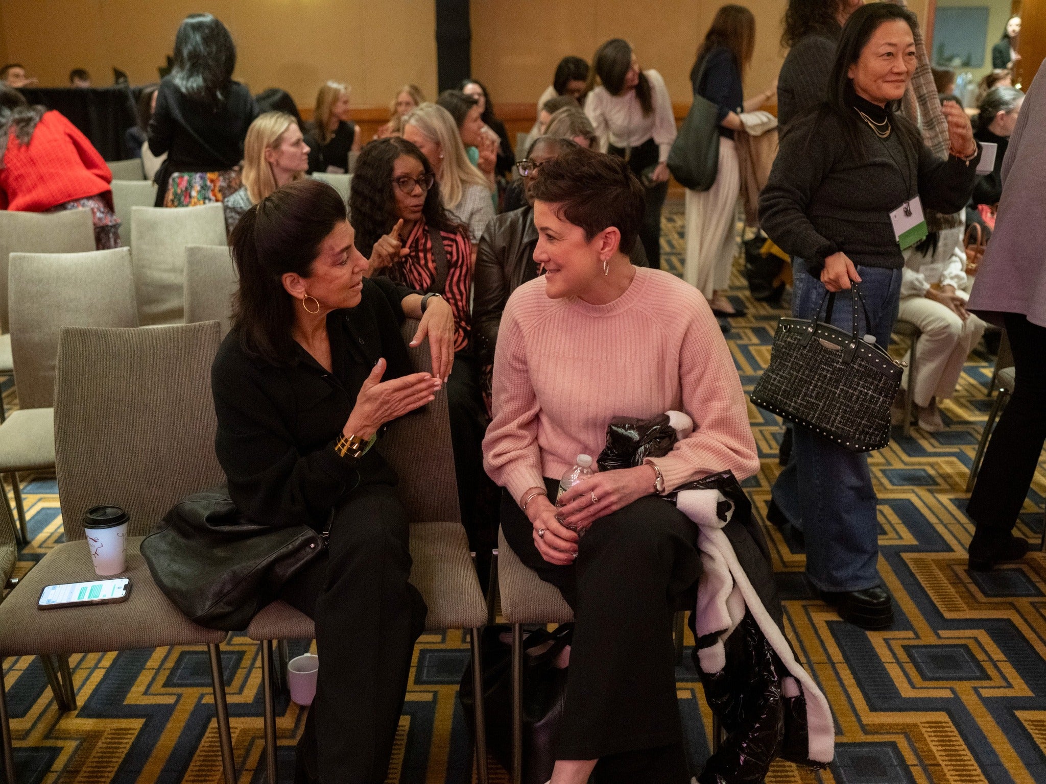 Two women sit in chairs at a conference session, engaged in animated conversation, while other participants stand and talk in the background.