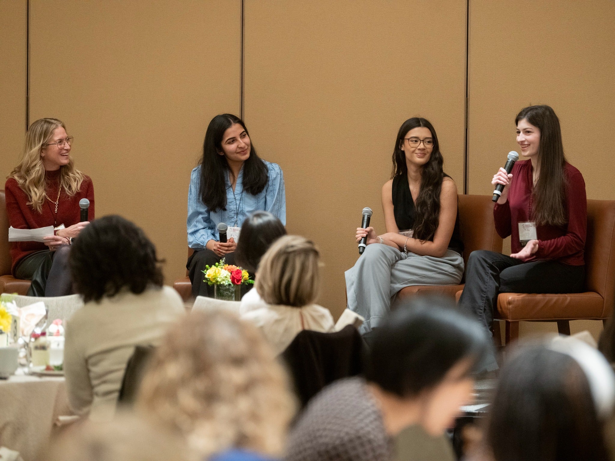 Four women sit on a low stage holding microphones, speaking as a panel to an audience seated at round tables in front of them.