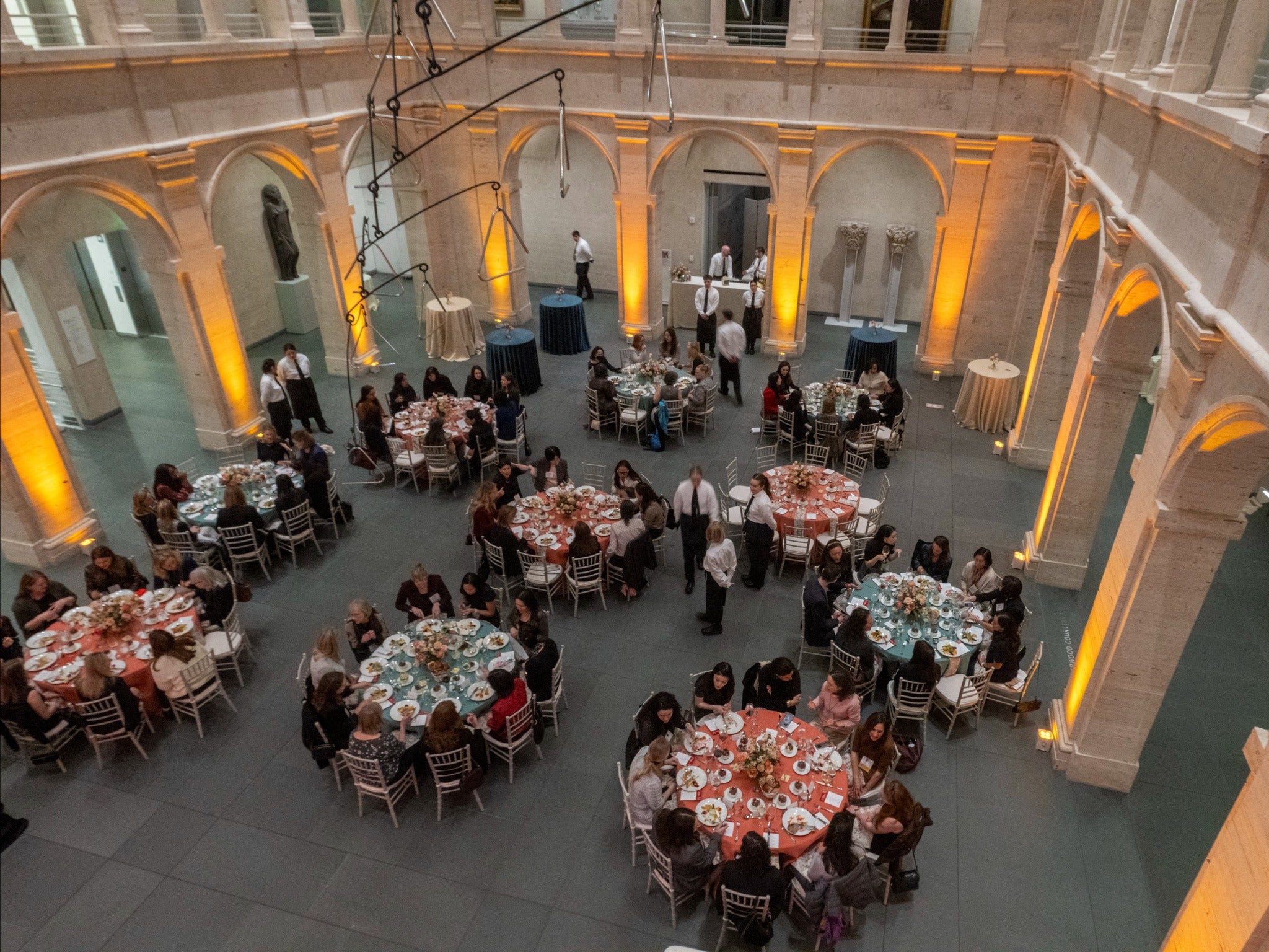 Overhead view of a grand hall with tall arches and glowing orange uplighting, where many round tables are set for dinner and filled with attendees.