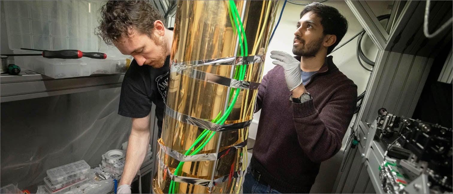 Two researchers working on either side of a large cylindrical piece of equipment wrapped in reflective gold material and bright green cables