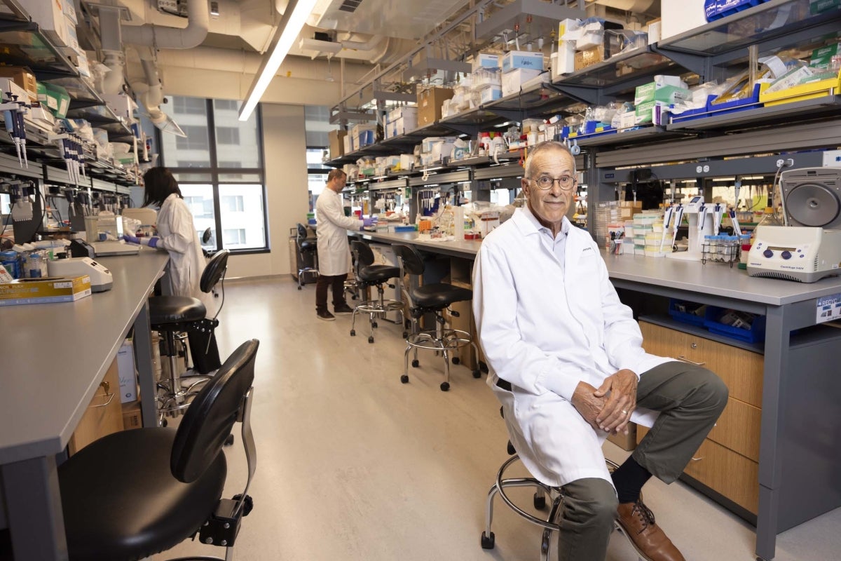 Harvard scientist David Walt in a white lab coat is sitting on a stool in a well-equipped laboratory. Behind him, shelves are filled with various lab equipment and supplies. Two other scientists in lab coats are working at benches in the background. The lab has bright lighting and large windows.