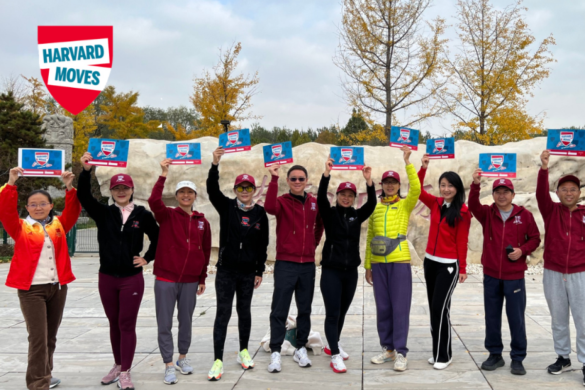 A group of Harvard alumni in Harvard gear during a past Harvard Moves event, holding up certificates. They stand before autumn trees and rock formations, showcasing the spirit of community and fitness.