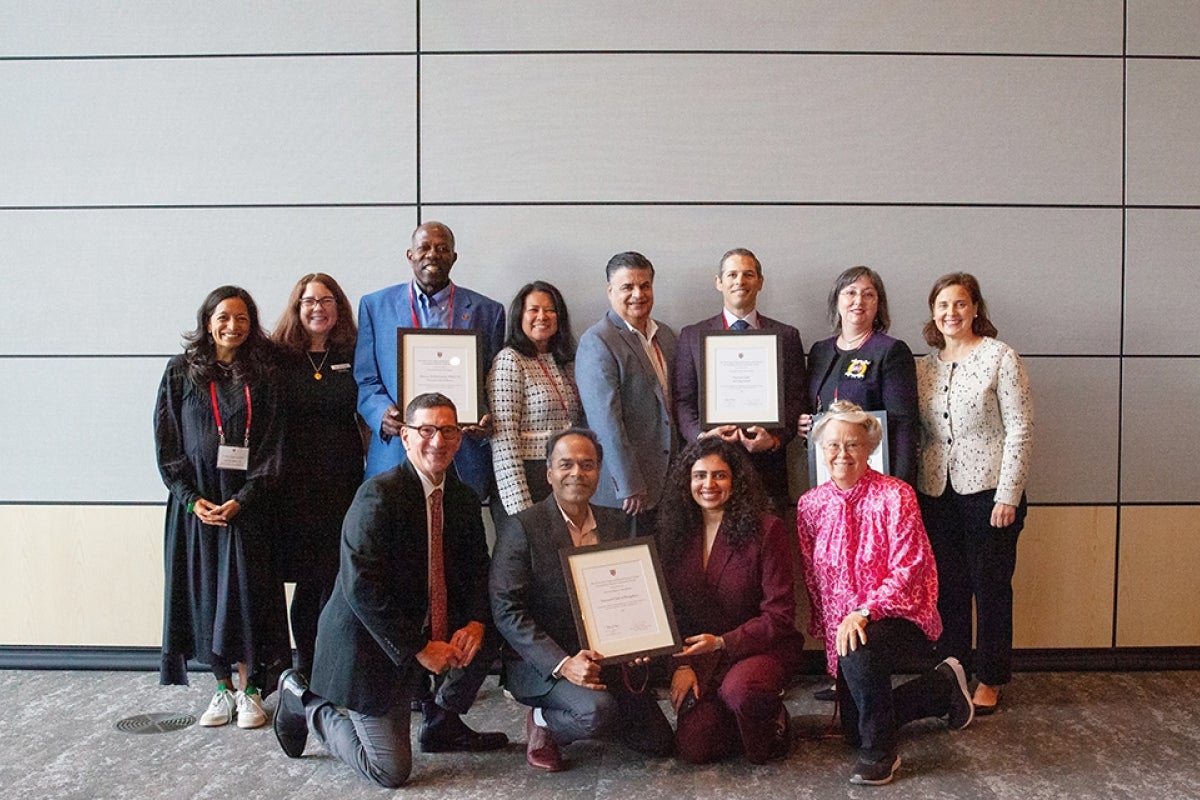 12 people pose for a group photo in front of gray wall with four people holding awards