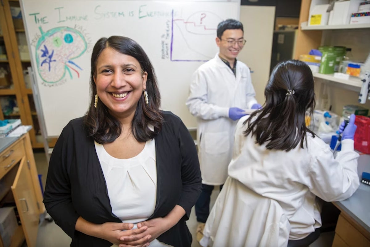 Girija Goyal stands in front of a whiteboard in her lab as two colleagues in white lab coats work in the background