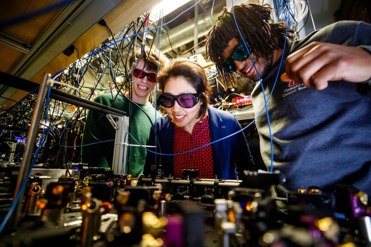 Three researchers wearing protective glasses examine a complex array of lenses and lasers in a lab.