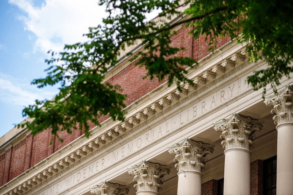 Close-up view of the front facade of a classical building made of red brick and stone. Prominent white columns with ornate Corinthian capitals support the entablature, which is engraved with the words “WIDENER MEMORIAL LIBRARY.” Green tree branches partially frame the top of the image against a partly cloudy sky.