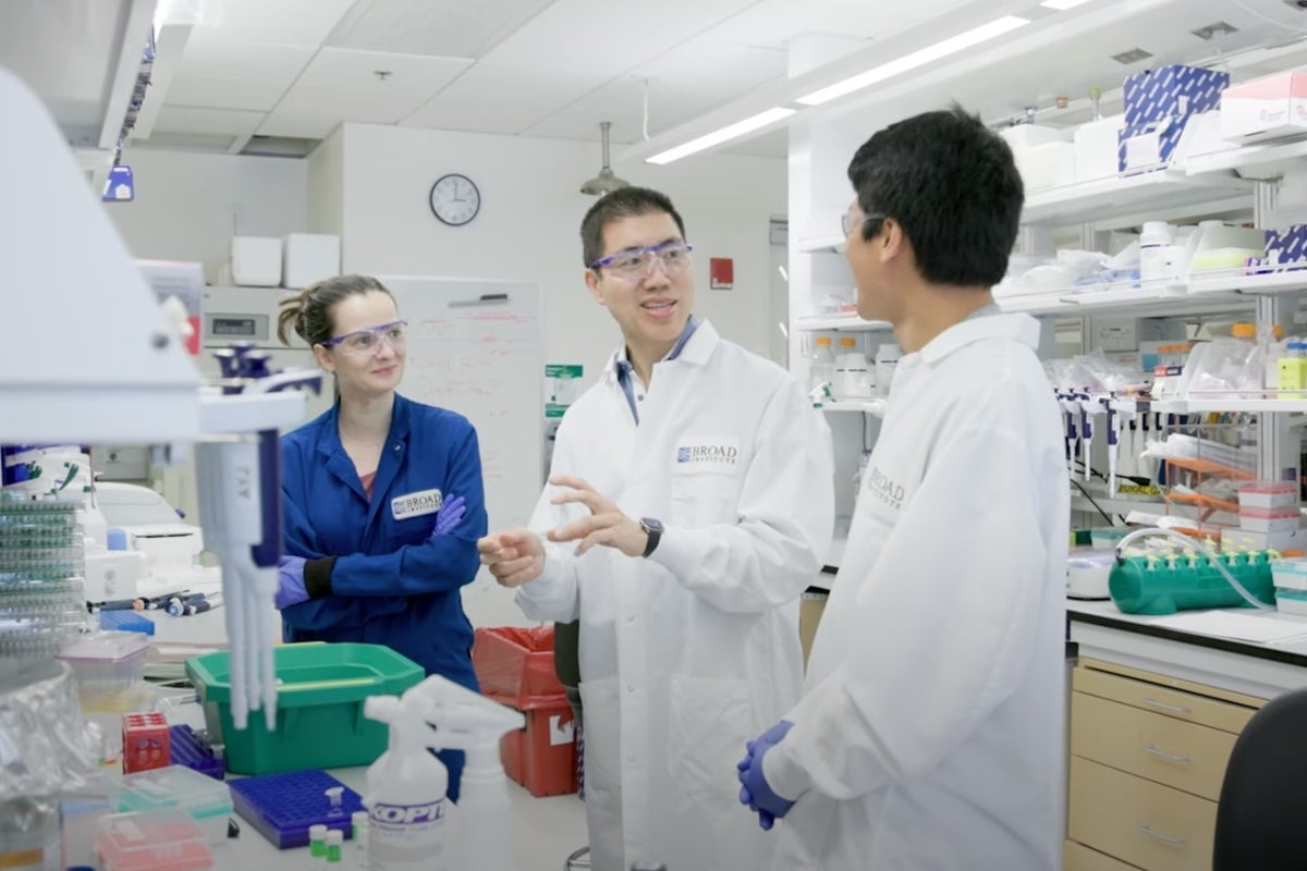 Three researchers wearing lab coats and safety goggles converse in a bright, modern laboratory at the Broad Institute. They stand surrounded by lab equipment, pipettes, and trays, engaged in discussion amid an active research workspace.