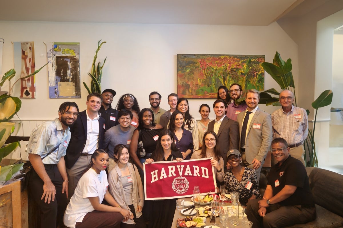 Group of recent Harvard alumni gathered indoors at a Welcome to Your City event, smiling and posing together with a crimson Harvard banner in front.