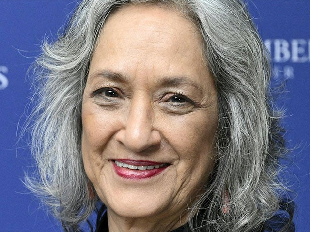 A smiling woman with shoulder-length gray hair poses against a blue backdrop.
