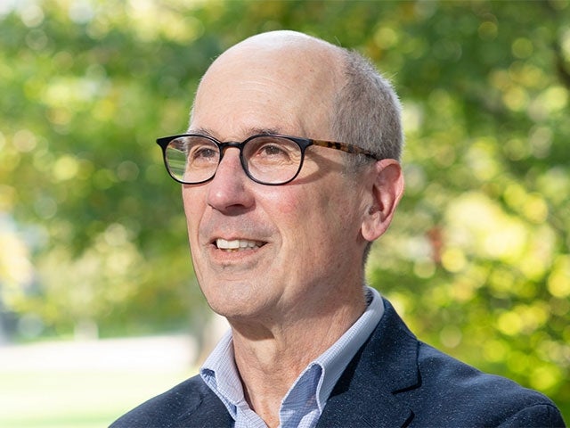 A smiling man wearing a suit stands in front of a tree on a sunny day.