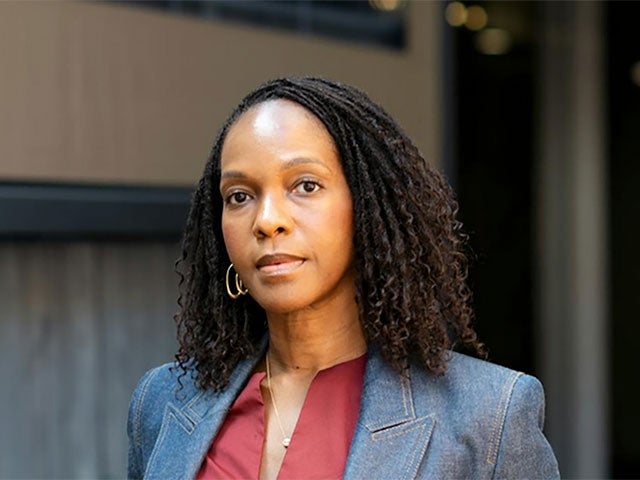 A woman with shoulder-length curly hair stands confidently with hands in her pockets, wearing a red dress and denim blazer, in an urban outdoor setting with neutral tones.