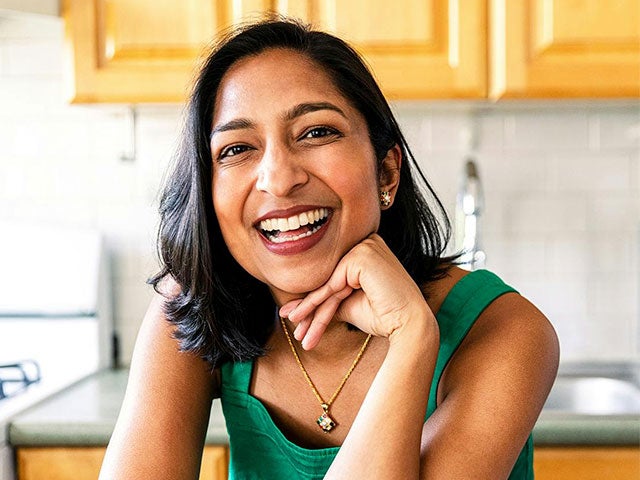 A smiling woman with shoulder-length dark hair rests her chin on her hand while seated at a kitchen counter, wearing a green sleeveless top and a gold necklace.