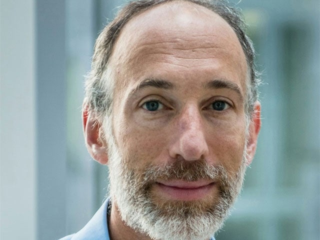 A man with a close-cropped beard and thinning hair looks directly at the camera, wearing a light blue shirt with a soft-focus indoor background.