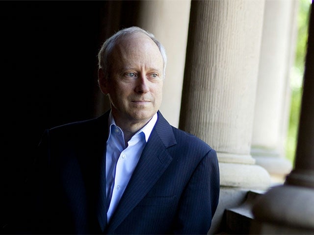 A man in a dark suit and light blue shirt stands pensively by a row of classical stone columns, gazing into the distance with soft natural light illuminating the scene.