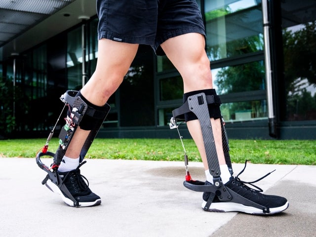 Person walking outdoors wearing robotic exoskeleton braces on both legs.