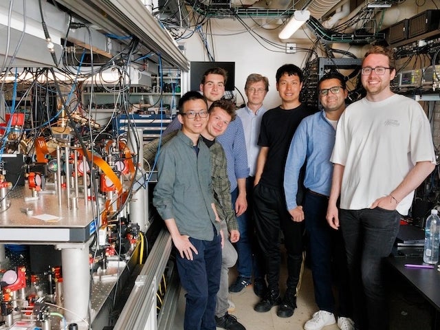 A group of scientists wear casual clothes in a lab surrounded by equipment and wiring