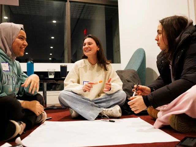 Three students sit in a circle smiling holding markers with a white paper.