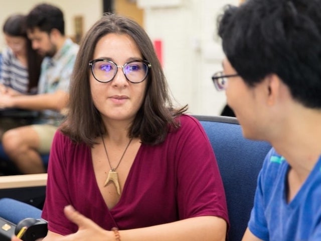 Two students speak to each other in a classroom setting while other students work at their desks in the background