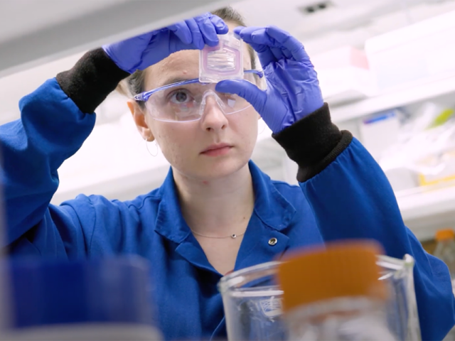 Scientist examines a small lab container while wearing safety goggles and gloves.