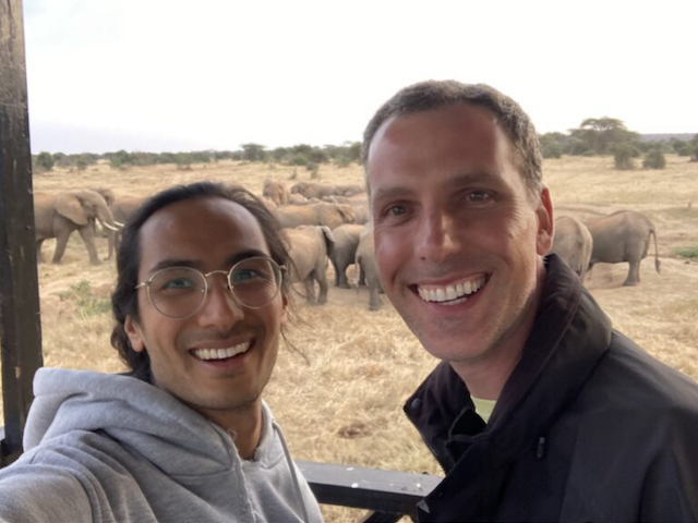 Viraj Sikand and Mark Tracy in a field with elephants in the background