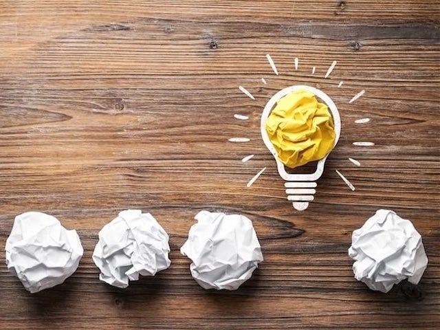 Four crumpled paper balls sit on a wooden table, with one yellow paper ball inside a drawn light bulb