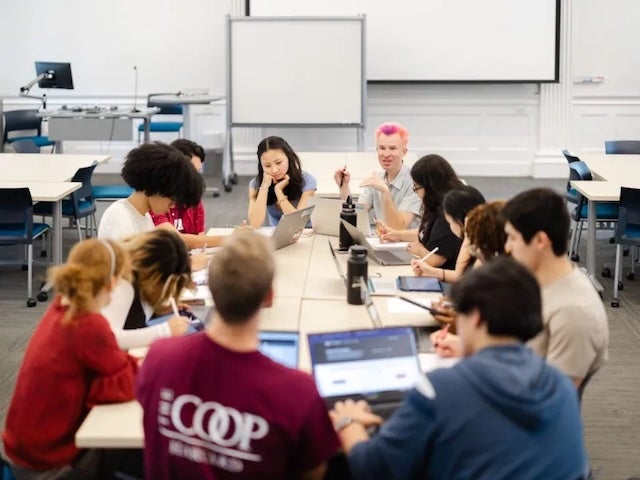  Students gathered around a table in a classroom, working on laptops and taking notes