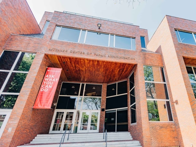 Exterior view of the Littauer Center for Public Administration at Harvard Kennedy School, featuring a prominent red banner reading "ASK WHAT YOU CAN DO" above the main entrance. 