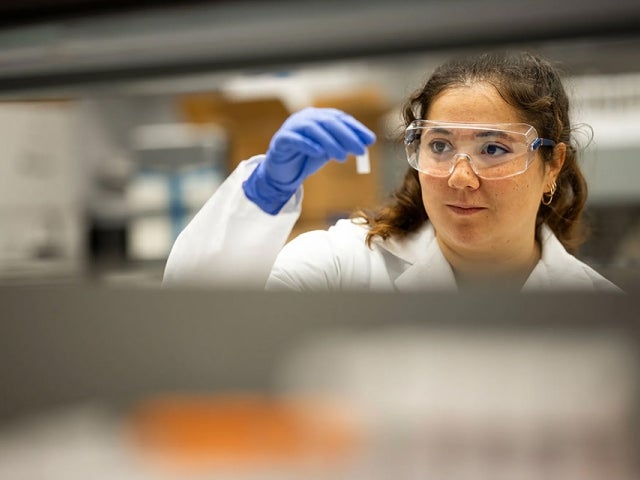 Olivia Hogan-Lopez in a lab coat and safety goggles examines a small sample vial