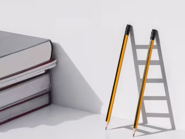 A stack of books next to two pencils leaning on a wall leaving the shadow of a ladder