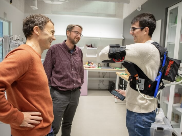 A researcher in a lab demonstrates the functionality of a soft, wearable robotic device while two other researchers look on.