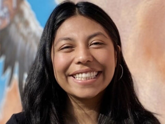 Karina Herrera smiles in front of a colorful mural with soft earthy tones and a blue sky. 