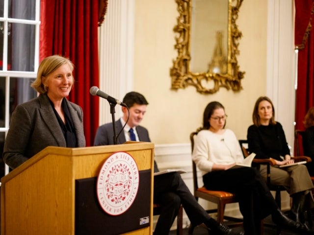 Hopi Hoekstra stands at a podium with a microphone, smiling as she addresses an audience. Behind her, four people are seated in chairs, holding papers, in a room with red curtains, a large window, and an ornate mirror on the wall.