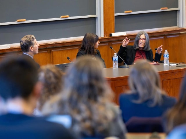 A panel of three speakers sits at the front of a classroom. Professor of Law Lucie White gestures while speaking, as the other two panelists listen. Several audience members sit in the foreground, facing the speakers.