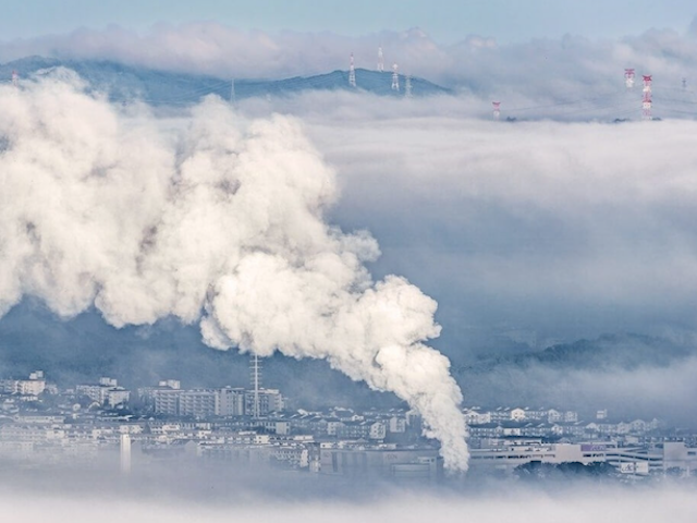 An industrial area is partially obscured by thick fog, with a prominent plume of white smoke rising into the air
