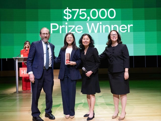 President Alan Garber on stage with PeriPeach founder Tess Kim, and Harvard i-lab managing director Becca Xiong and senior director Meagan Hall in front of a green-tiled screen displaying the words “$75,000 Prize Winner” 