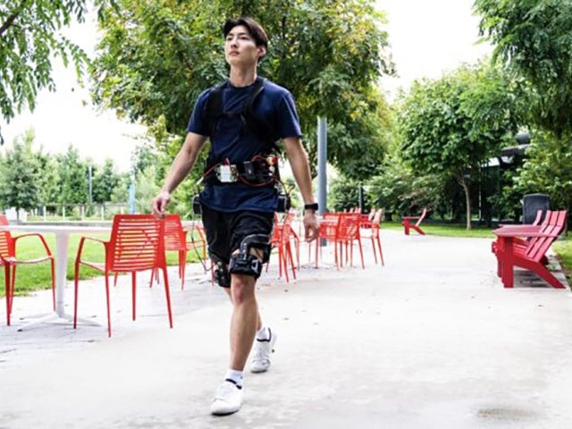 A person walks outdoors on a paved path wearing a prototype of an "exoskeleton.” Red chairs and tables are scattered along the path, with green trees and grass surrounding the area.