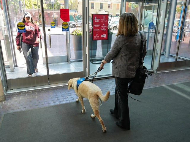 A person walks through glass doors with a service dog on a leash as another person holds the door open.
