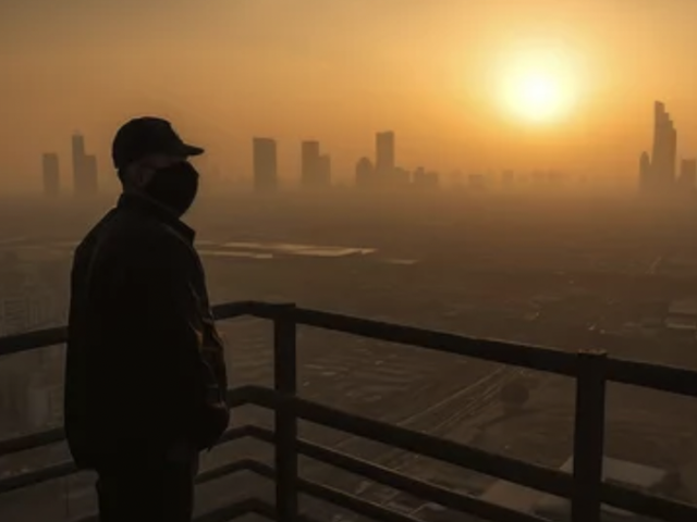 A man wearing a medical mask stands on a balcony overlooking a hazy smog-filled city skyline