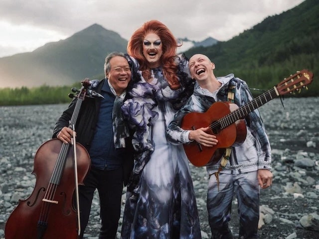 Yo-Yo Ma holding a cello, Pattie Gonia, and Quinn Christopherson holding an acoustic guitar standing outside with mountains in the background