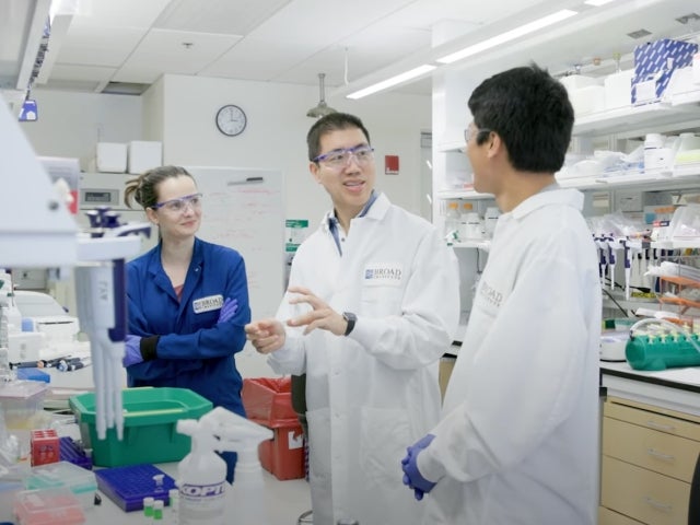 Three researchers wearing lab coats and safety goggles converse in a bright, modern laboratory at the Broad Institute. They stand surrounded by lab equipment, pipettes, and trays, engaged in discussion amid an active research workspace.