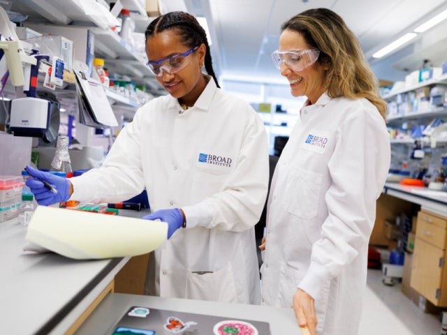 Two researchers wearing lab coats and safety goggles review notes together at a laboratory bench.