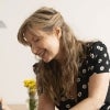 A woman wearing a short-sleeved dress is smiling while fixing an electronic device on a table.