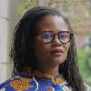 A woman with long locs and tortoiseshell glasses stands confidently in a colorful patterned top, with a blurred outdoor background of greenery and architecture.