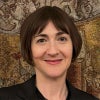 A smiling woman with short brown hair wears a black blazer and pendant necklace, posed in front of an intricately patterned tapestry.