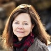 A woman with long brown hair and blue eyes gazes softly at the camera, wearing a dark scarf with red accents and a textured brown jacket, with a blurred outdoor background.