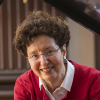 A smiling woman, wearing glasses, earrings, collared shirt, and sweater, leans on a wooden piano.